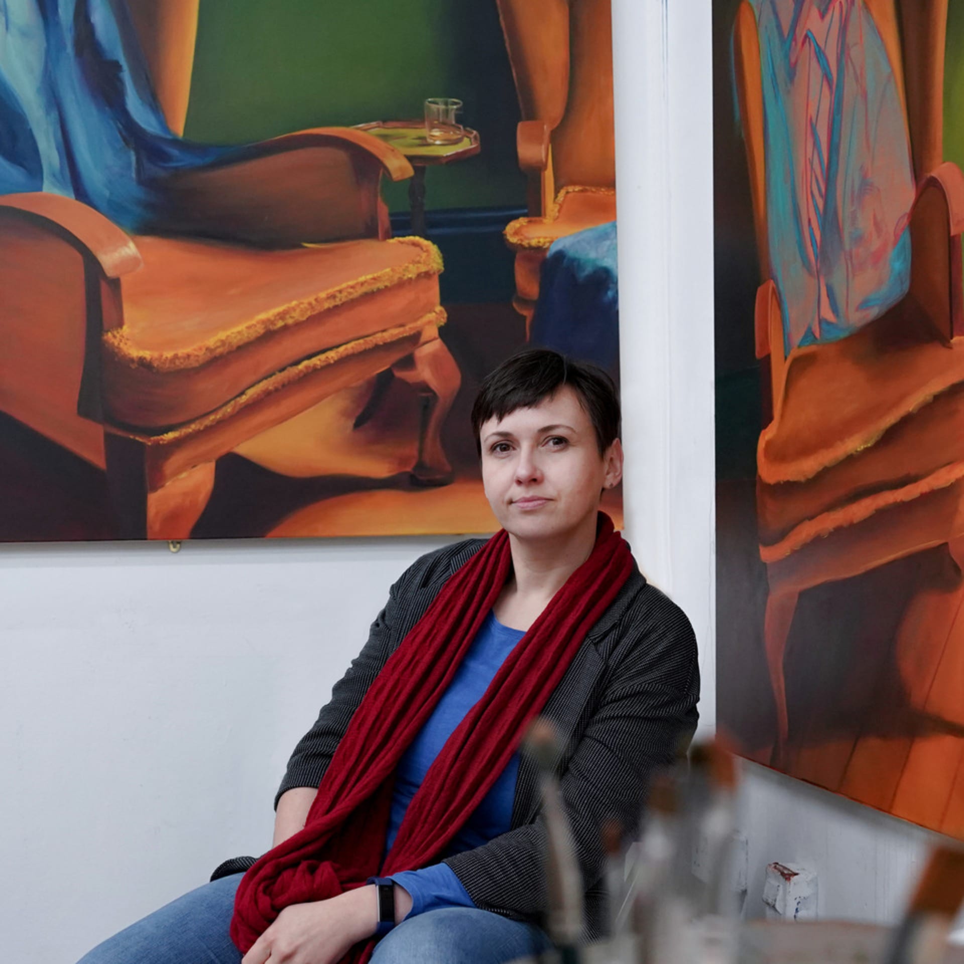 Portrait of the artist Susanne Baumann in front of two of her oil paintings. In the foreground are a few unsharp brushes.