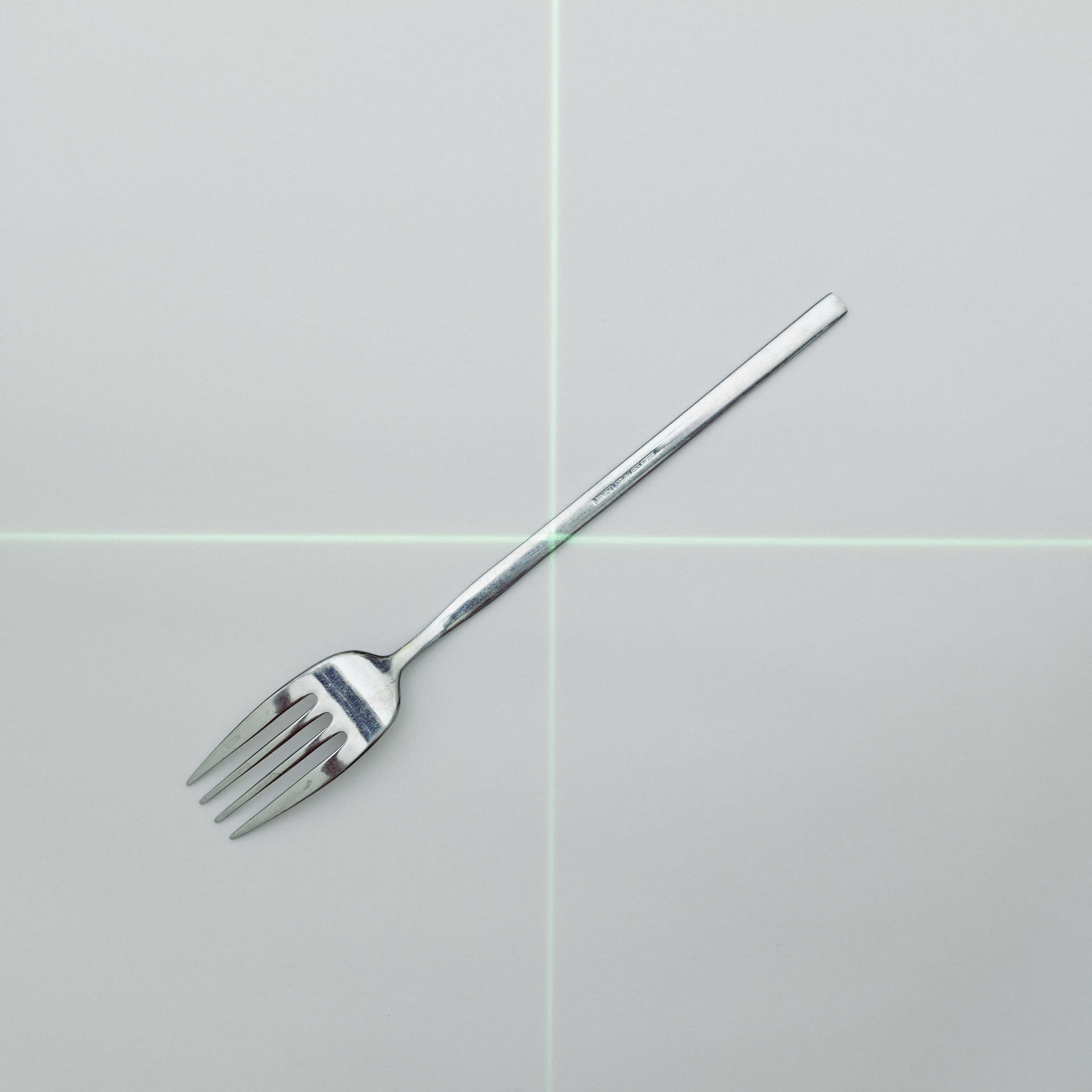 A green cross-shaped laser is projected onto a metallic fork against a pure white background.