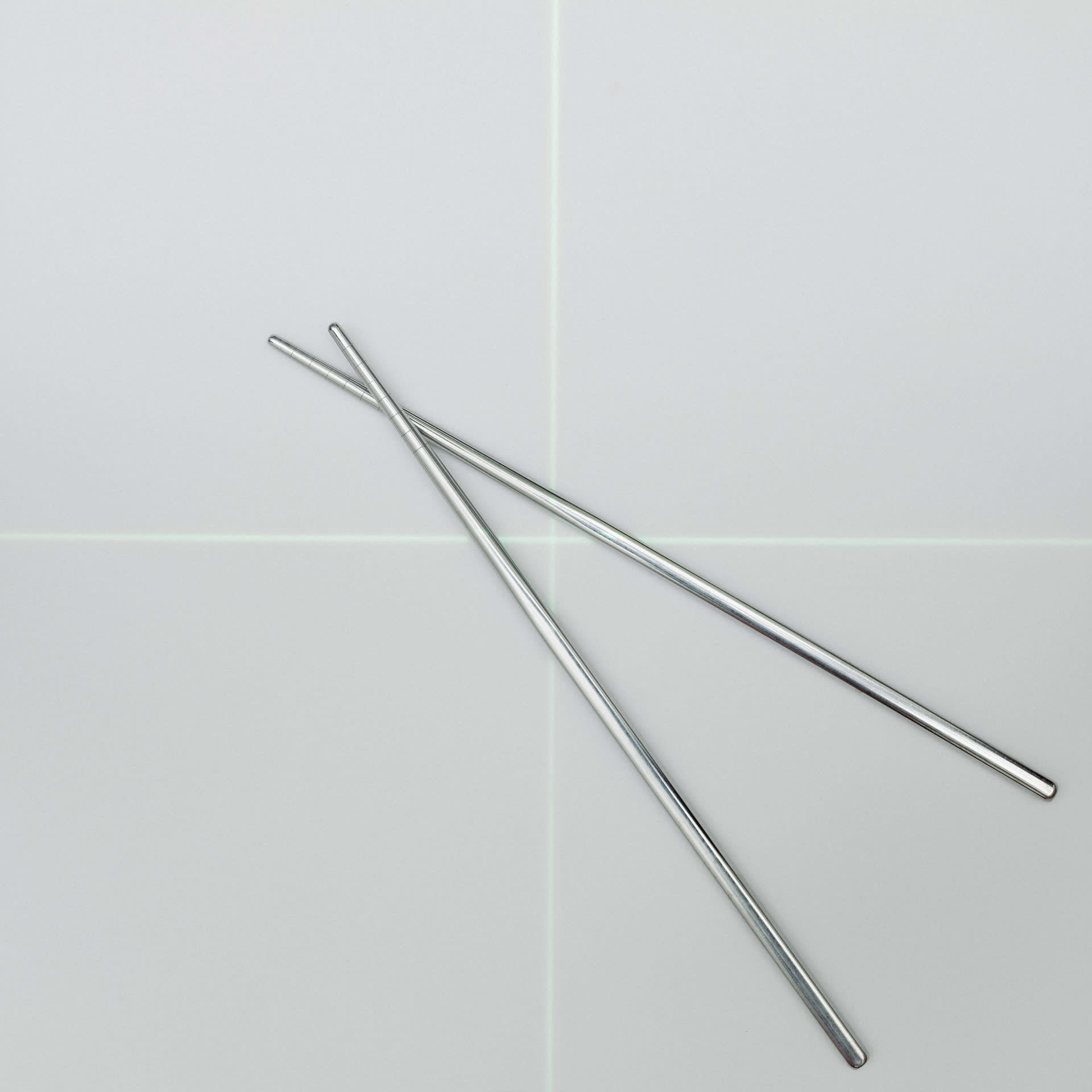A green cross-shaped laser is projected onto a metal chopstick against a pure white background.