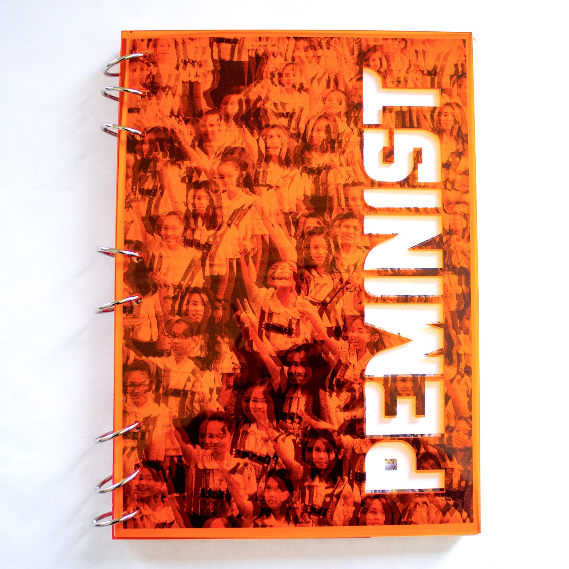 The image shows a book called 'Peminist,' with an orange acrylic cover and binded with silver rings/ 