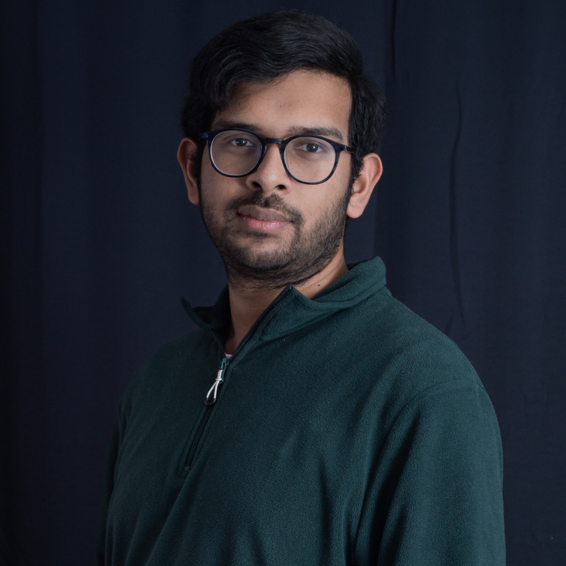 A south asian man with round glasses, full beard and a dark green jumper. 