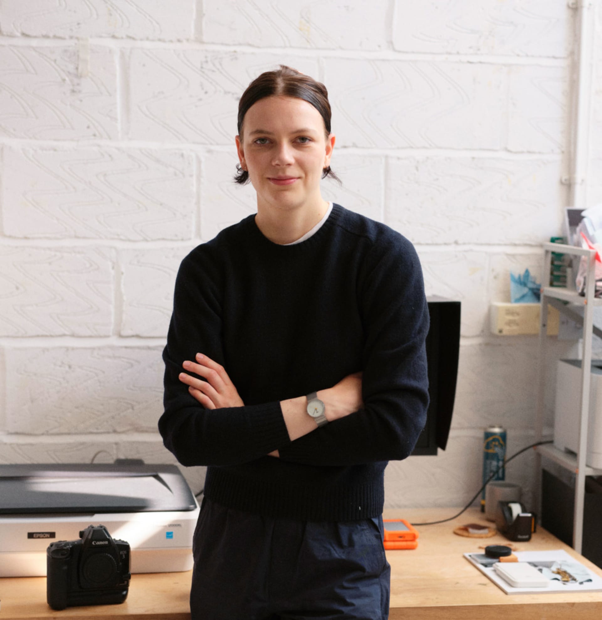 Portrait of Beatrice wearing a navy sweater, in a studio space, with a brick white background and working desk behind. 