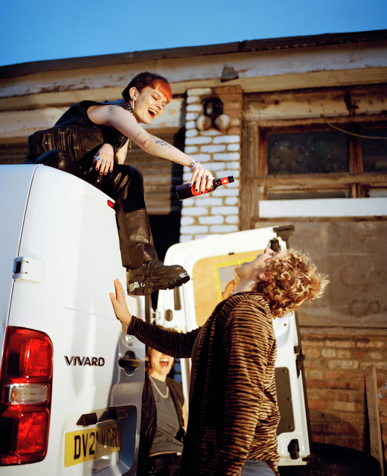 A photograph of a dyke sat on top of a white van and pouring beer into the mouth of a person below