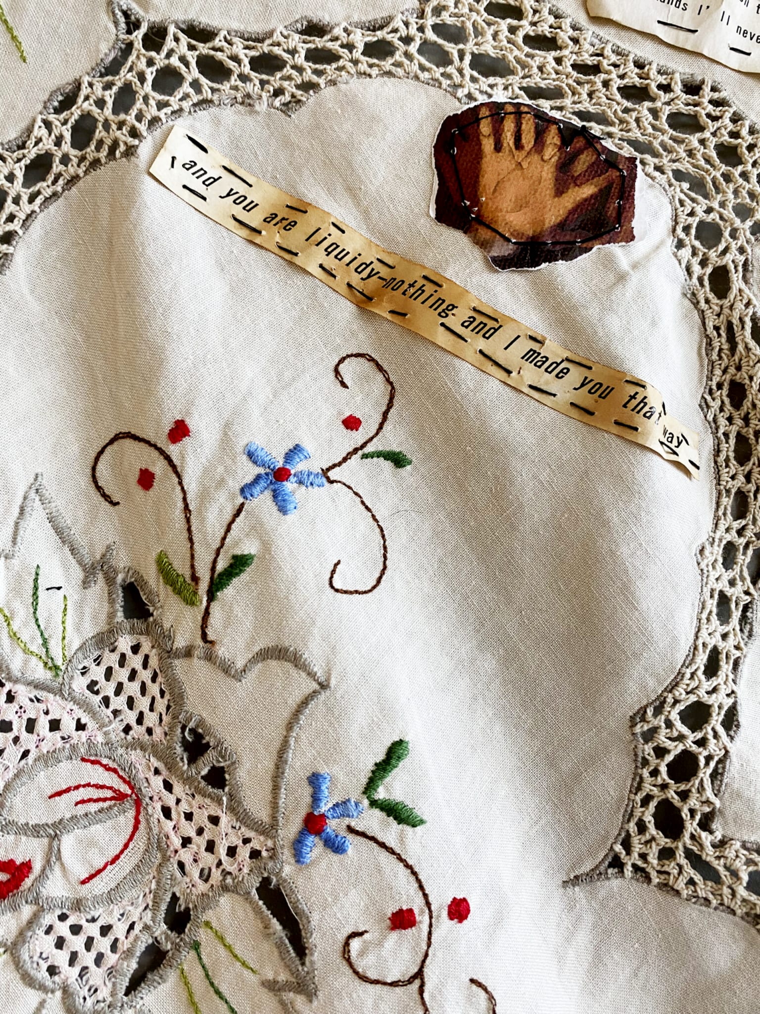 Detail of embroidered tablecloth with paper text stitched and image of hand stitched on (text not fully readable)