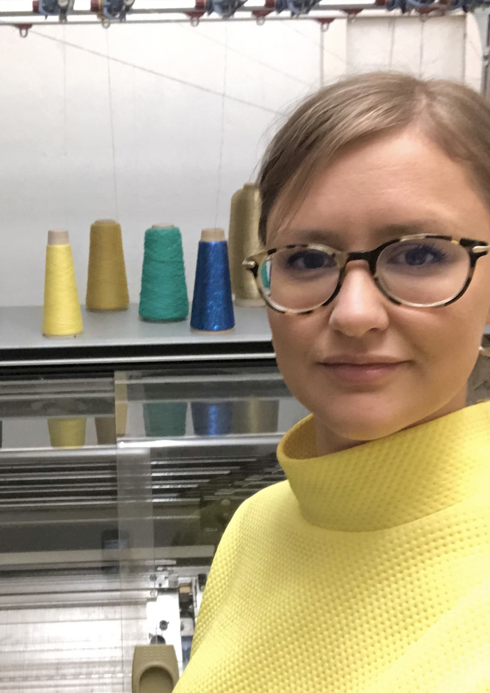 Cassie stands in front of colourful yarns on top of her Stoll knitting machine