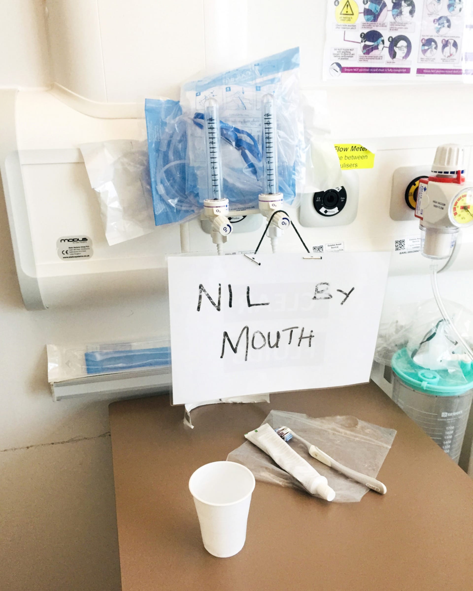 Photograph of a written sign reading, "NIL BY MOUTH" above a hospital side table with white plastic cup, toothpaste + toothbrush