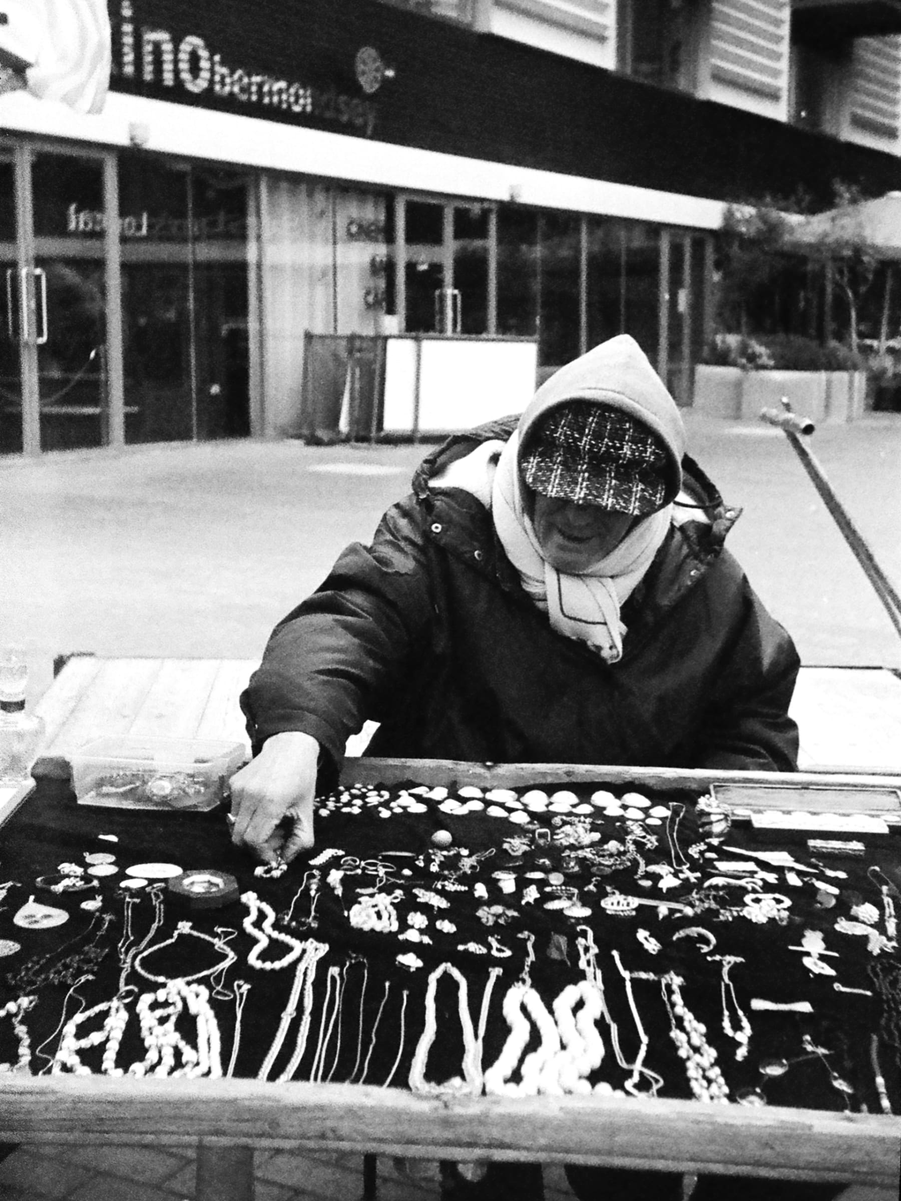 A photo of a man sat down behind his market stall with his hood up. his stall has jewellery which he reaches for to organise.