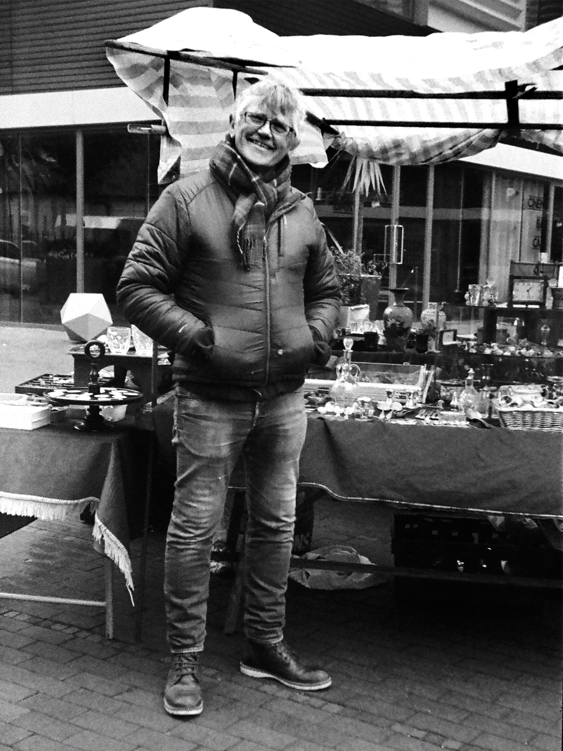 a man stands smiling on a market next to his stall and his wares to sell. He has a scarf on and puffy coat, it is winter. 