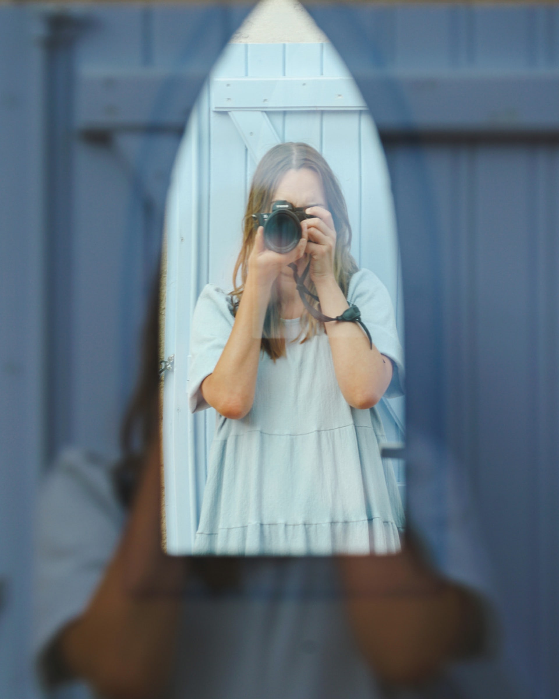 A woman wearing a blue dress is taking a photograph in a mirror behind a window creating a double reflection.