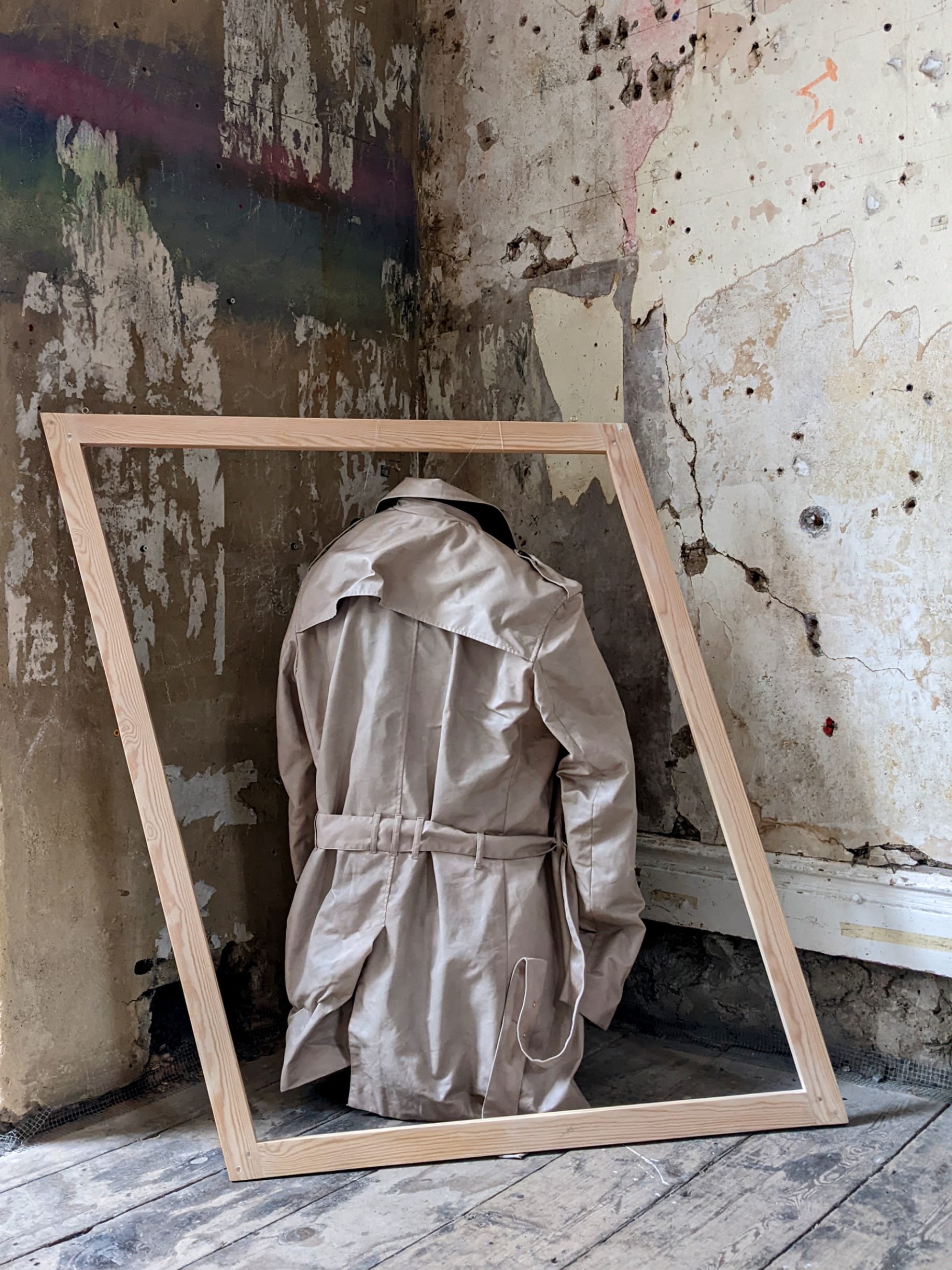 A hardened trenchcoat in an asymmetric wooden frame stands in the corner of a room, showing us his backside.