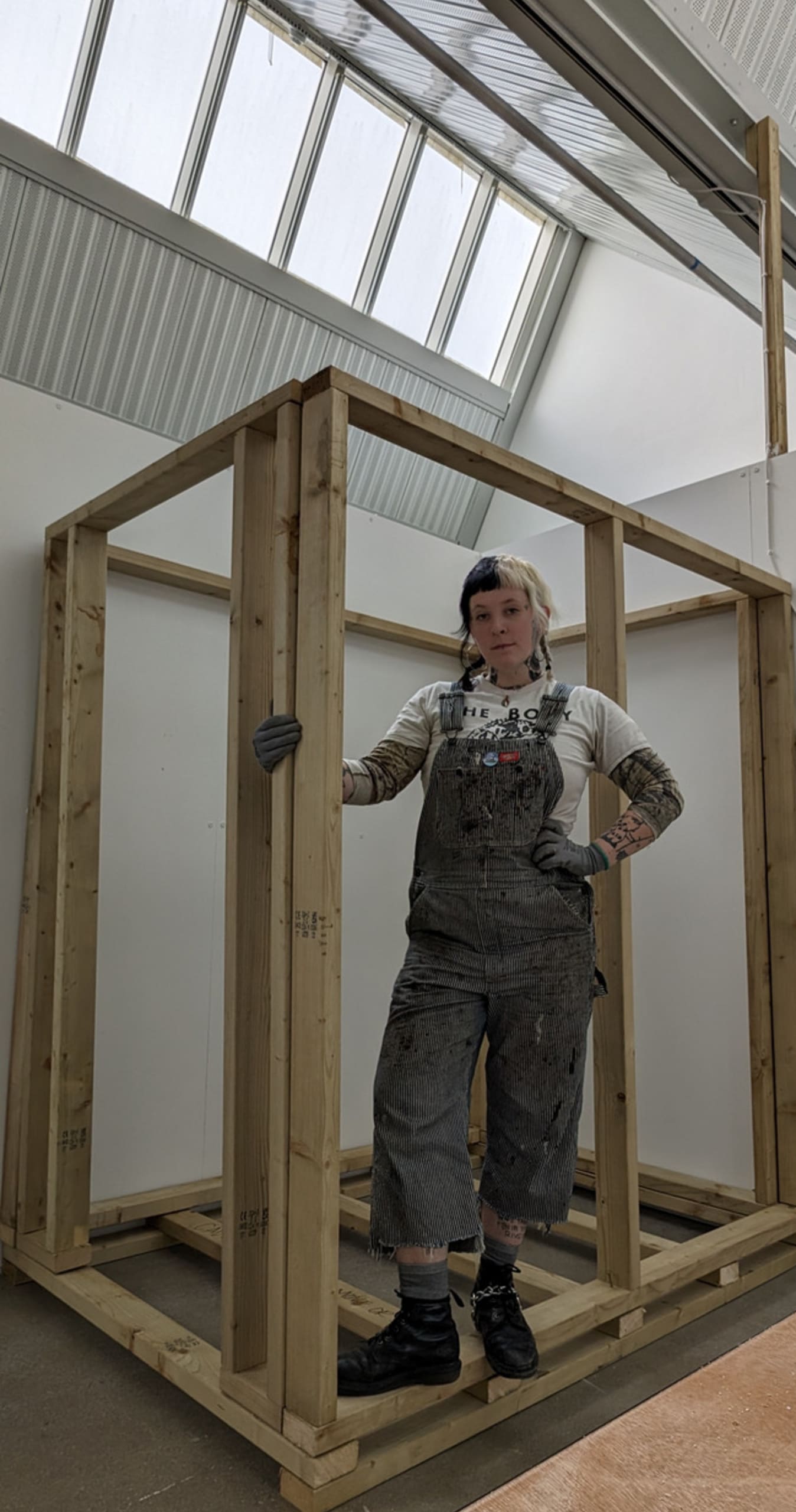 Person standing in overalls on wooden frame of a house. Person covered in tattoos and has split dye hair in plats. 