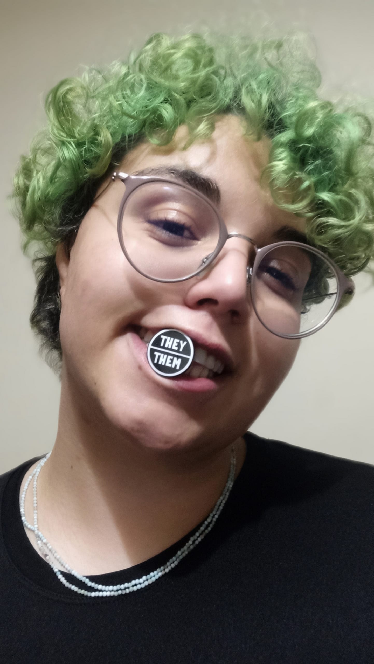 ID: Adri is smiling at the camera while holding a black and white 'they/them' pronouns pin between their teeth.