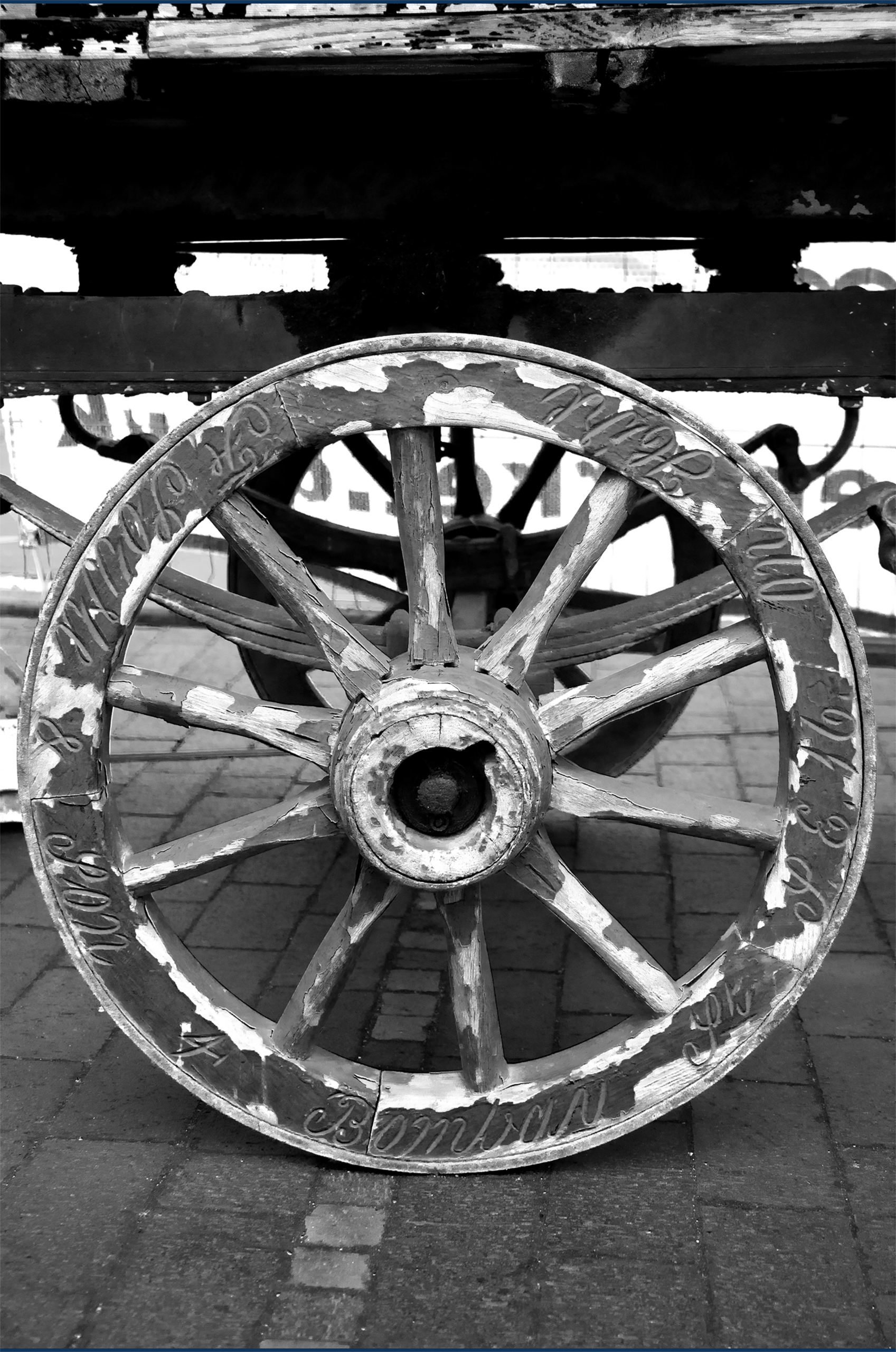 a photograph of a large and very old wagon wheel on cobblestones with cracking paint and names inscribed into its rim. 