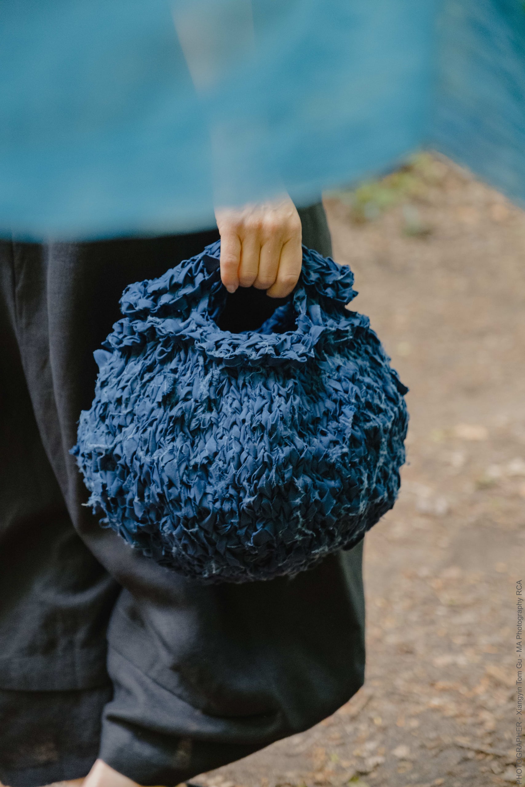 Round blue knitted fabric bag held by someone wearing black partially obscured by natural indigo dyed semi-transparent fabric