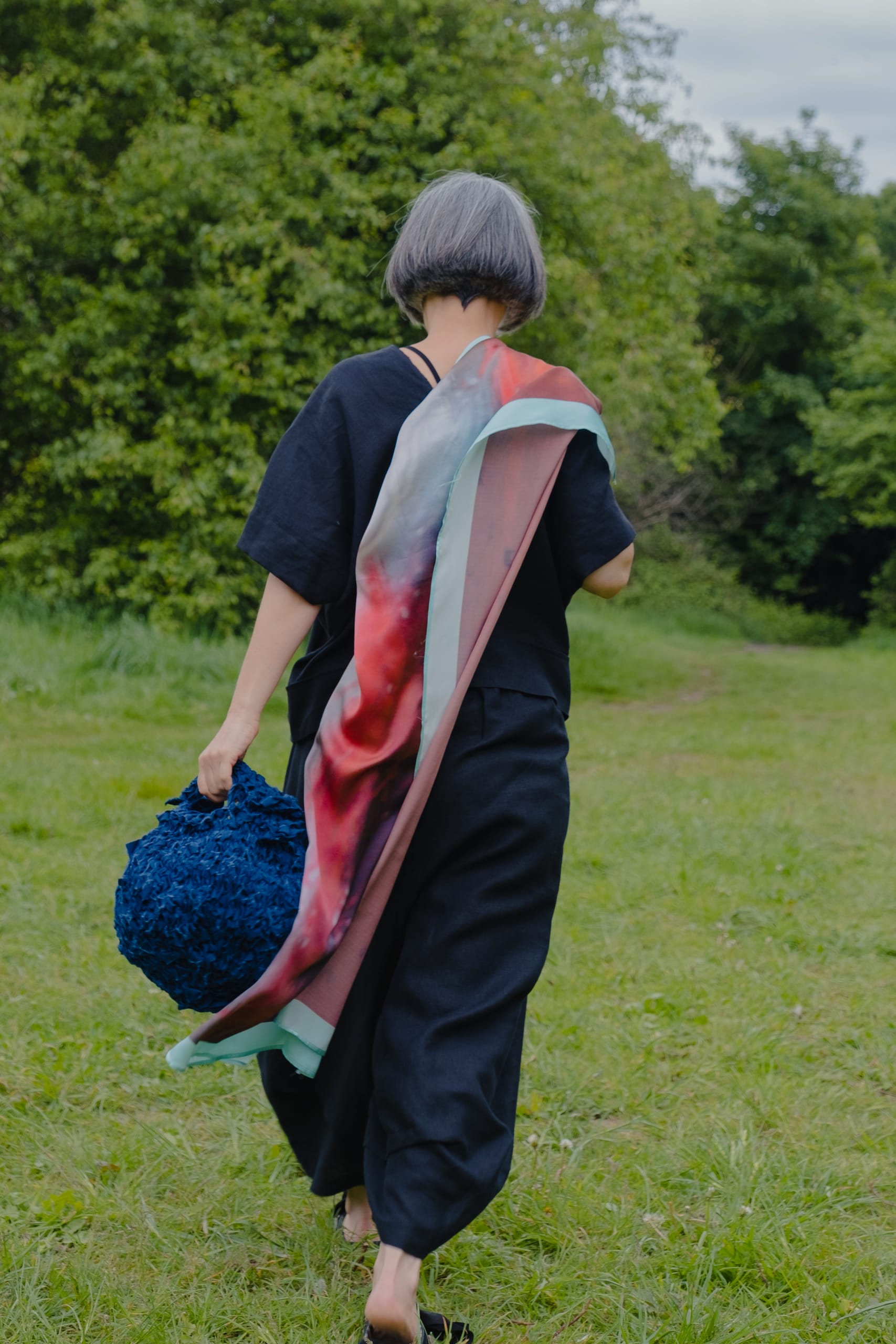 Walking away over the grass wearing 39.9 black linen trousers and top with red scarf over shoulder carrying blue mini sakk