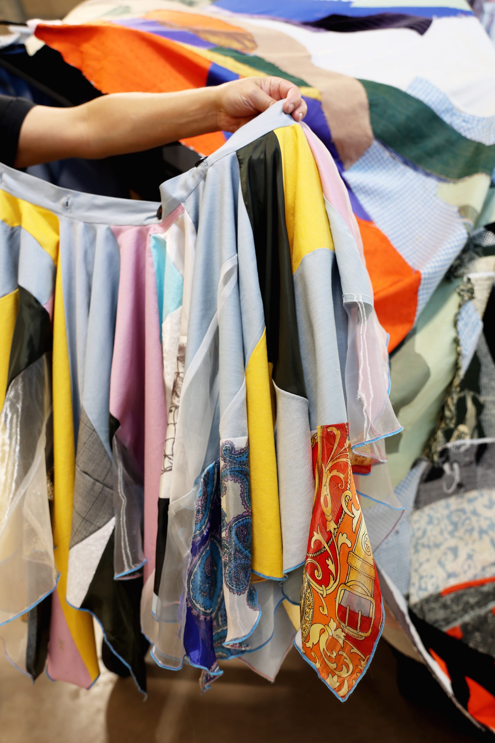 A hand is holding up a patchwork dress infront of a textile sculpture. 