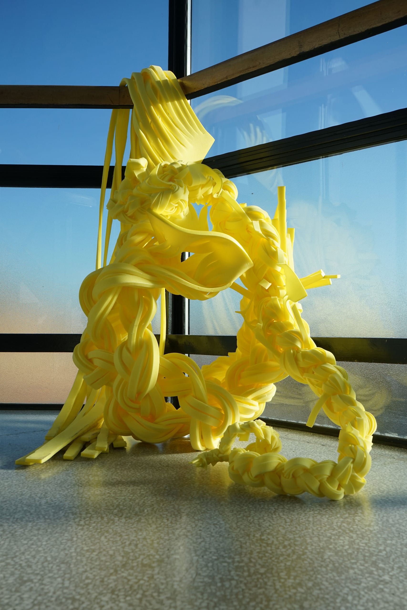 Braided yellow foam sculpture is cascading from a banister with a window as the backdrop where there is a blue sky