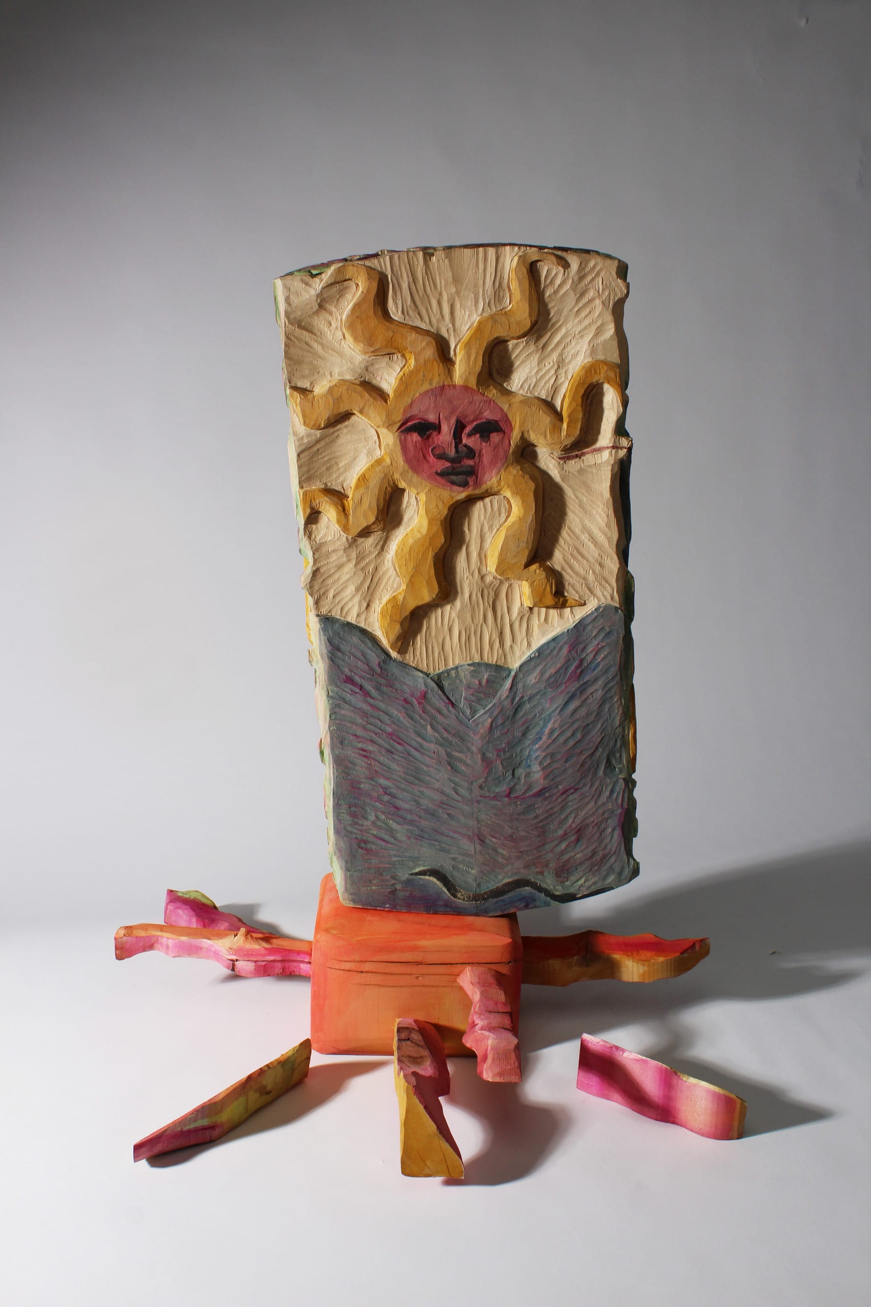 A carved sun protrudes out of a wooden block. They look on above watery mountains with a pink face and yellow coils.