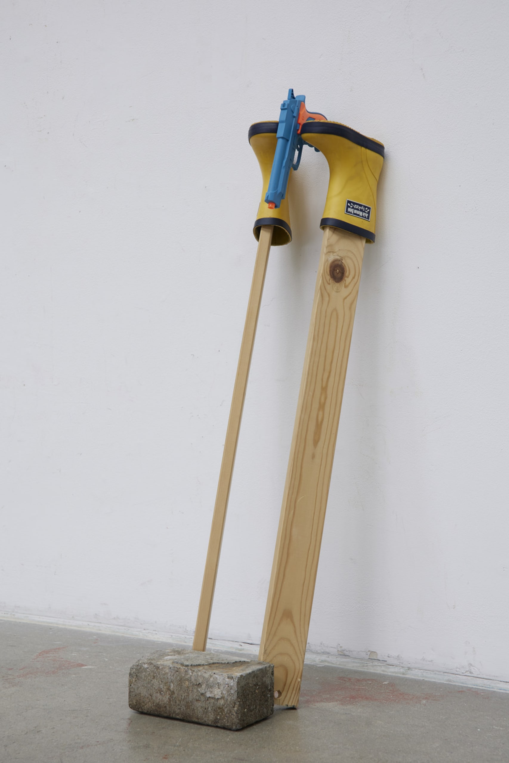 Photo of an assemblage sculpture leaning against a wall. It consists of a brick, two pieces of wood, kids boots, and a toy gun.