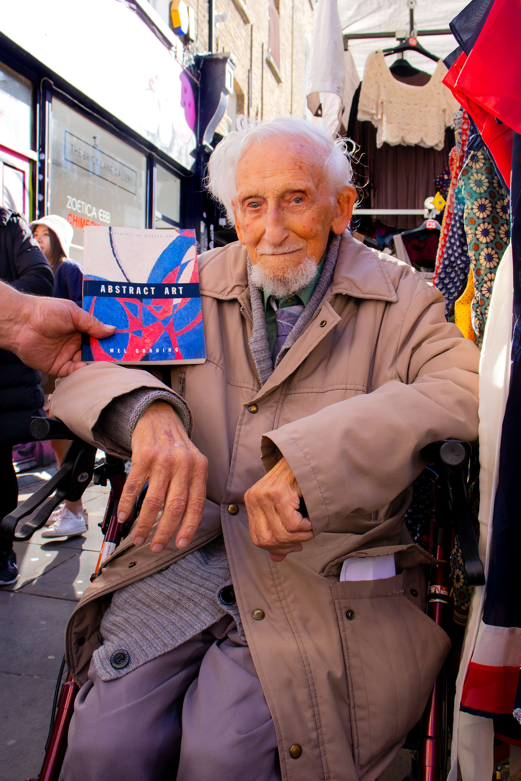 A elderly man in a wheelchair poses for a photo, he smiles, a man out of shot next to him holds up a book that says abstract art