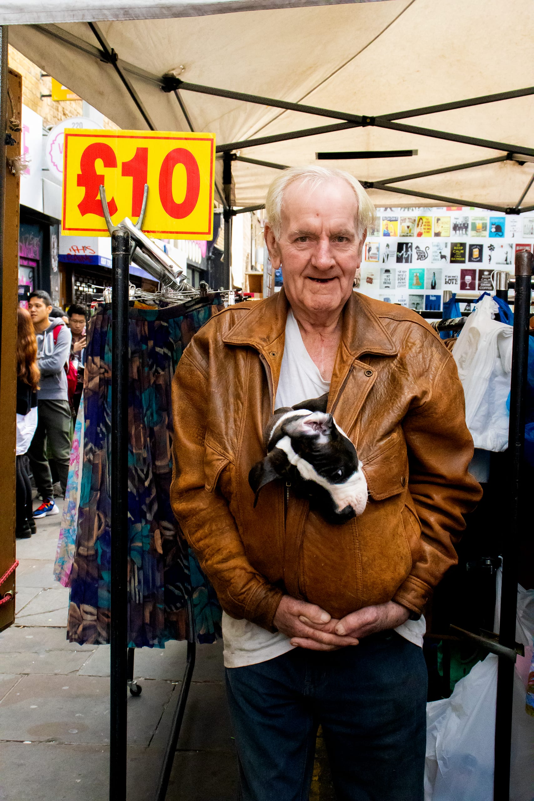 An older man poses in front of a clothing stall, he smiles, there is a small puppy with its head poking out of his jacket. 