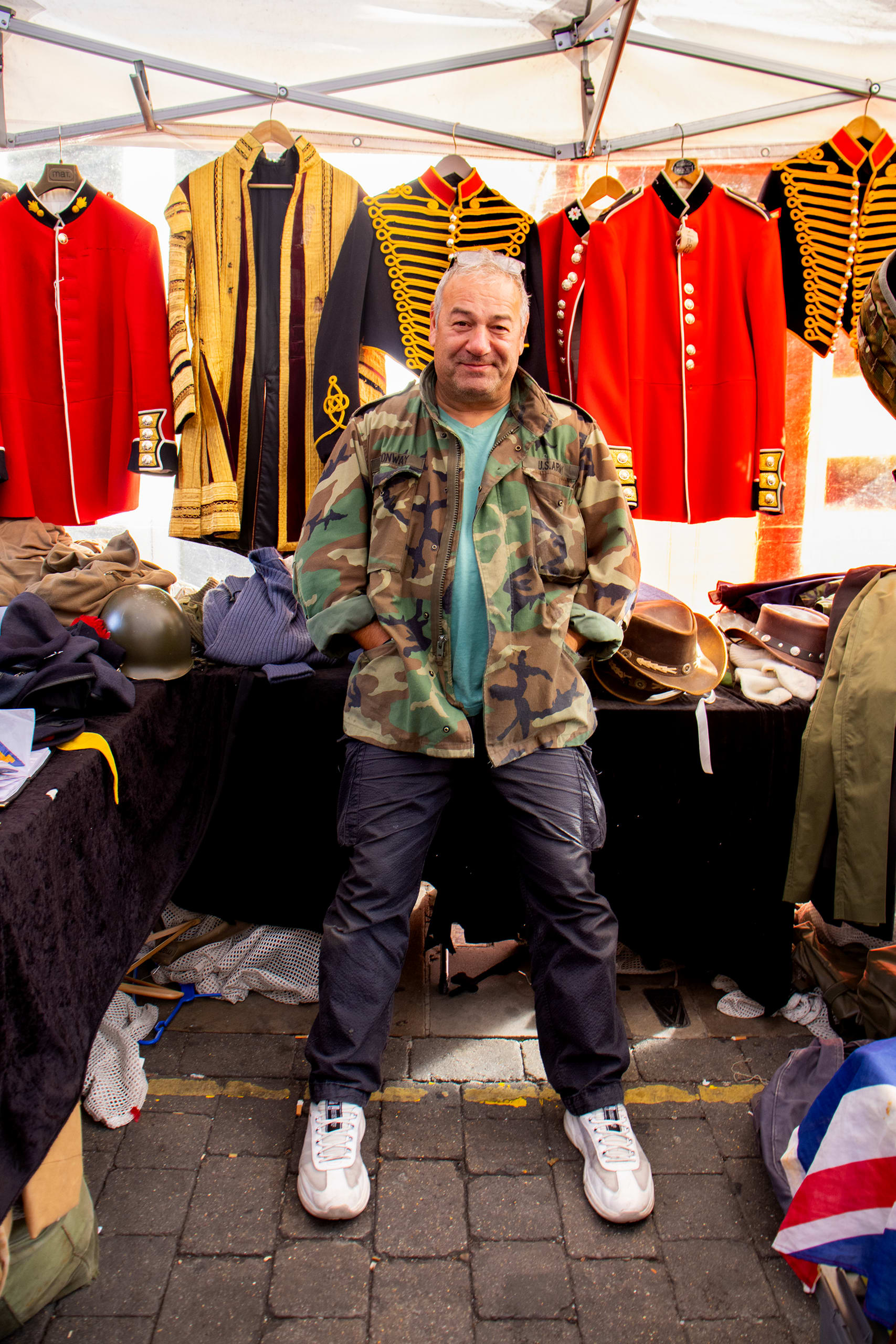 A man in camo clothing leans on his market stall looking at the camera, behind him is various fine antique militaria coats