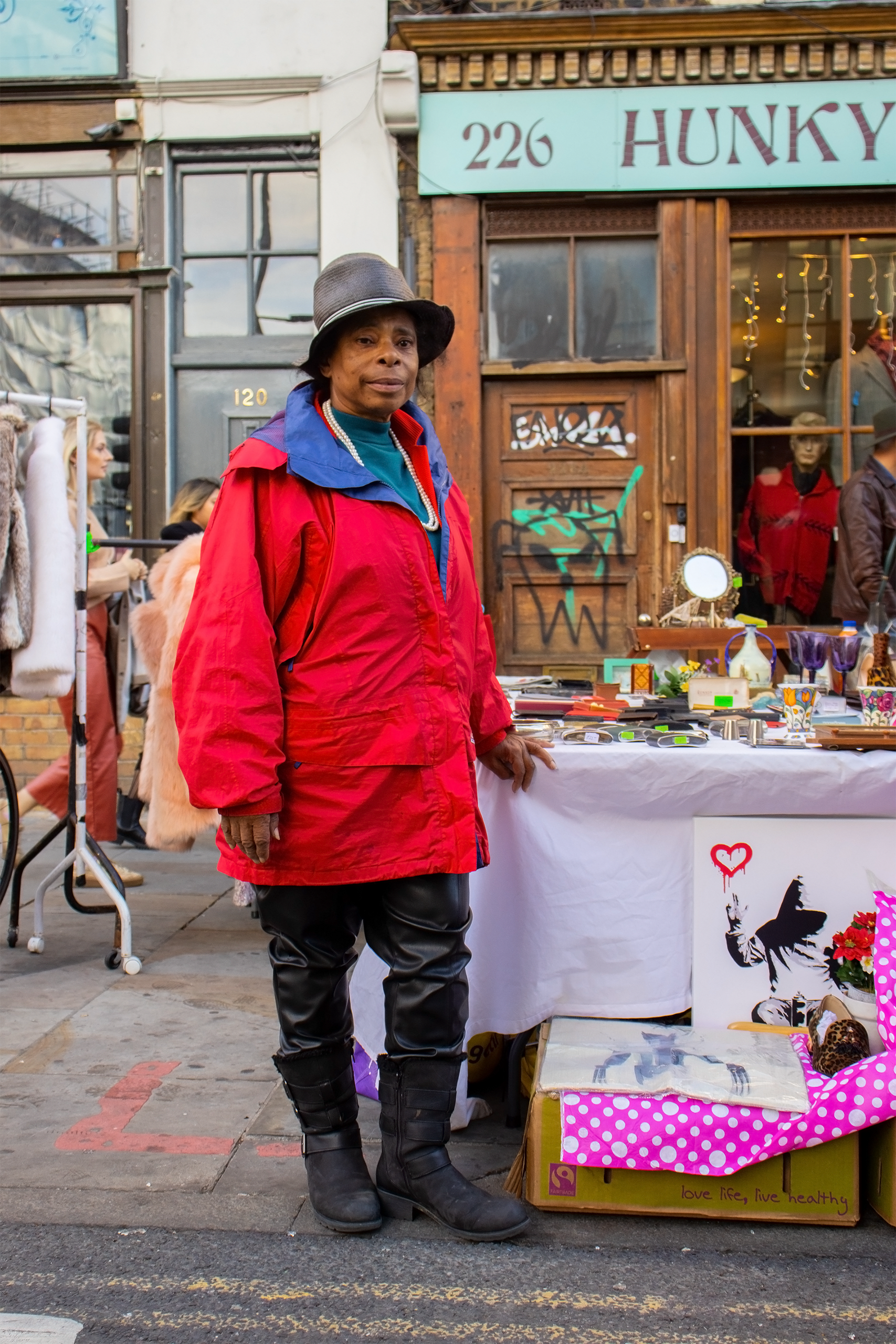 A black lady with a hat on looks out at the camera she is standing leaning on her stall looking pensive 