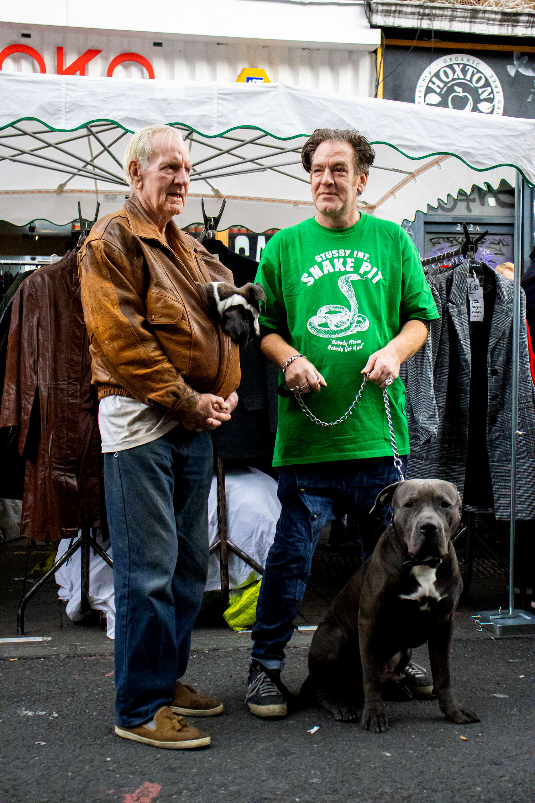 two men pose for a portrait they are father and son. The son has a very large dog on a chain lead the father has a small puppy