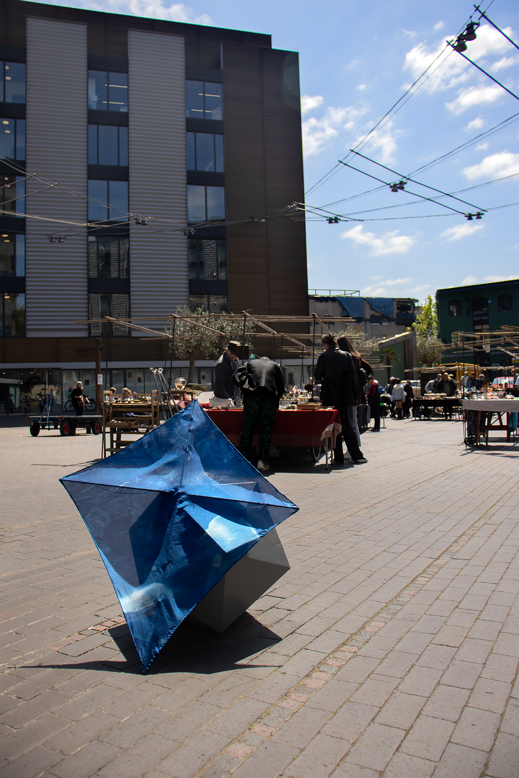 A kite sits in the foreground against a rock, with an antique market in the background, there are customers and it is sunny