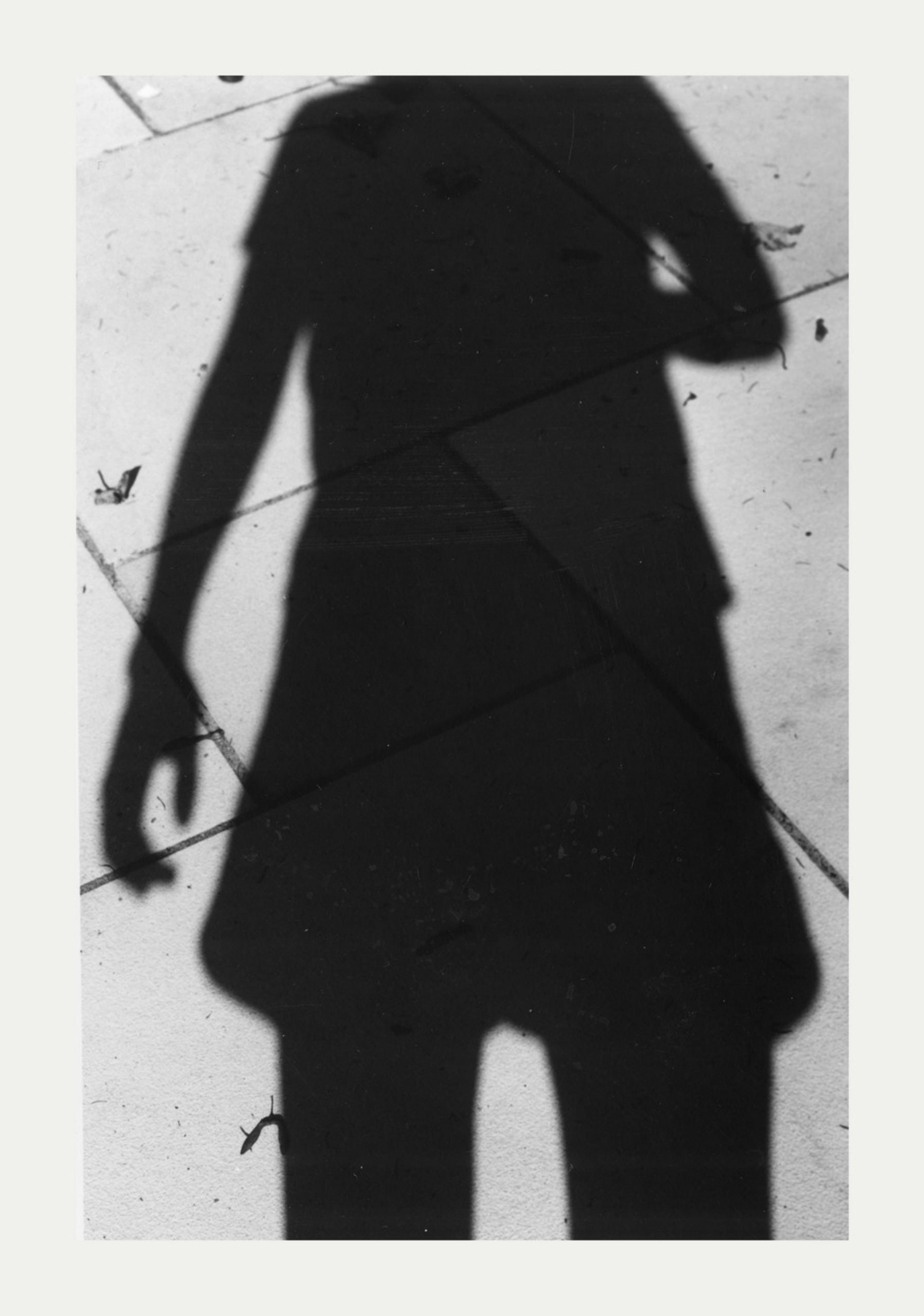 A black and white photographic print of a shadow on a tiled ground, with the head and feet of the figure missing from the image