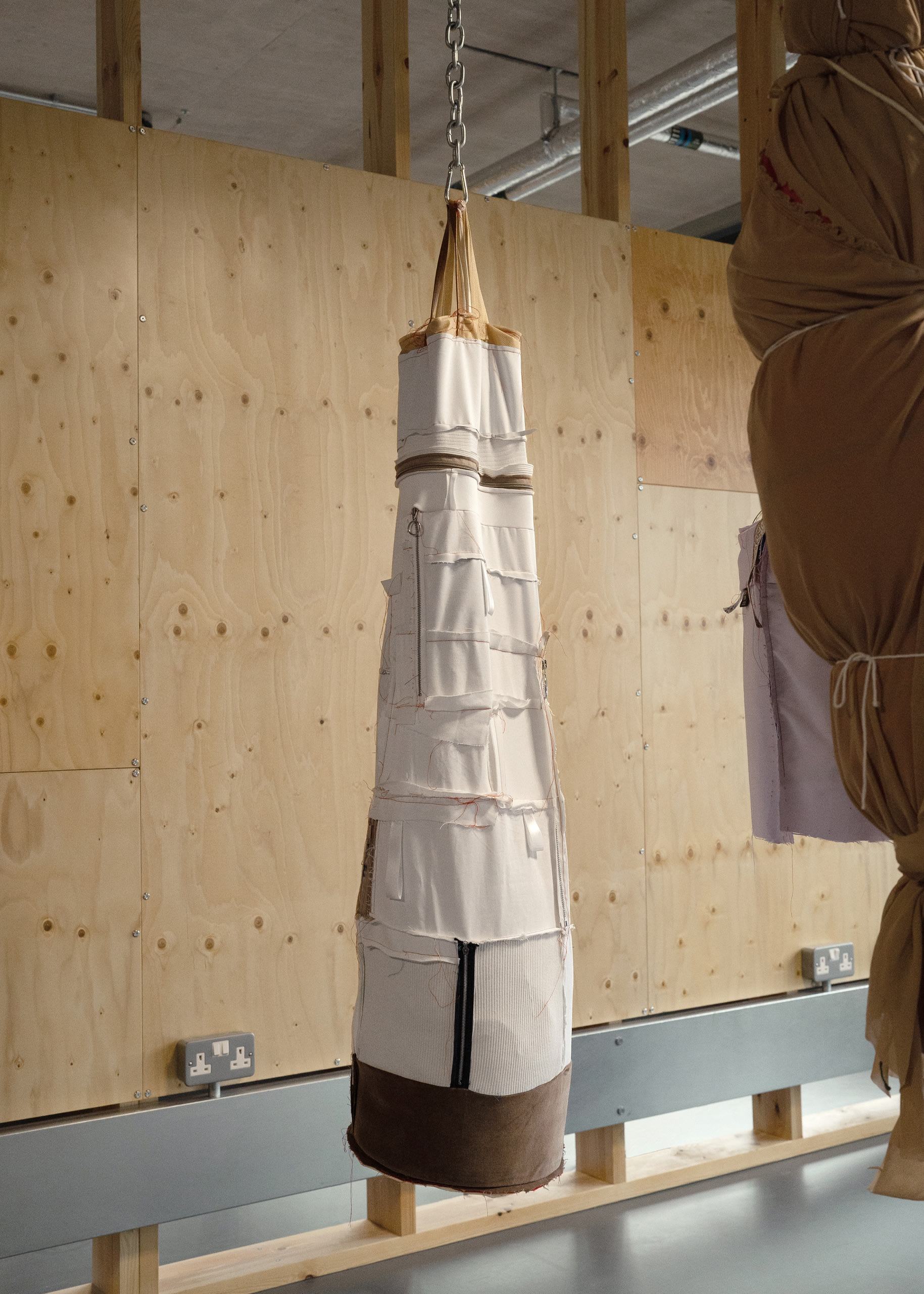 Object hanging from metal chain resembling a punch bag and made from scraps of white material with excessive number of zips