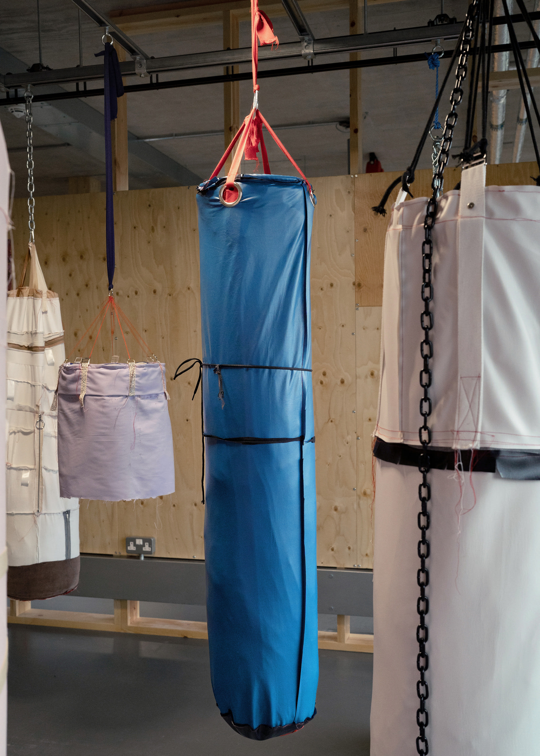 Blue leatherette cylindrical object hanging from ceiling by red webbing. Others hang in the periphery
