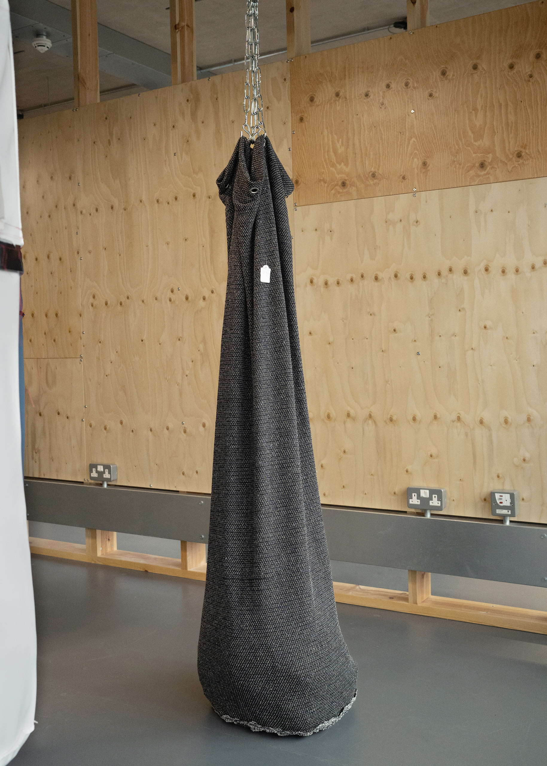 One sagging black object resembling a punch bag hanging by thick chain and resting on the floor with a shop tag protruding