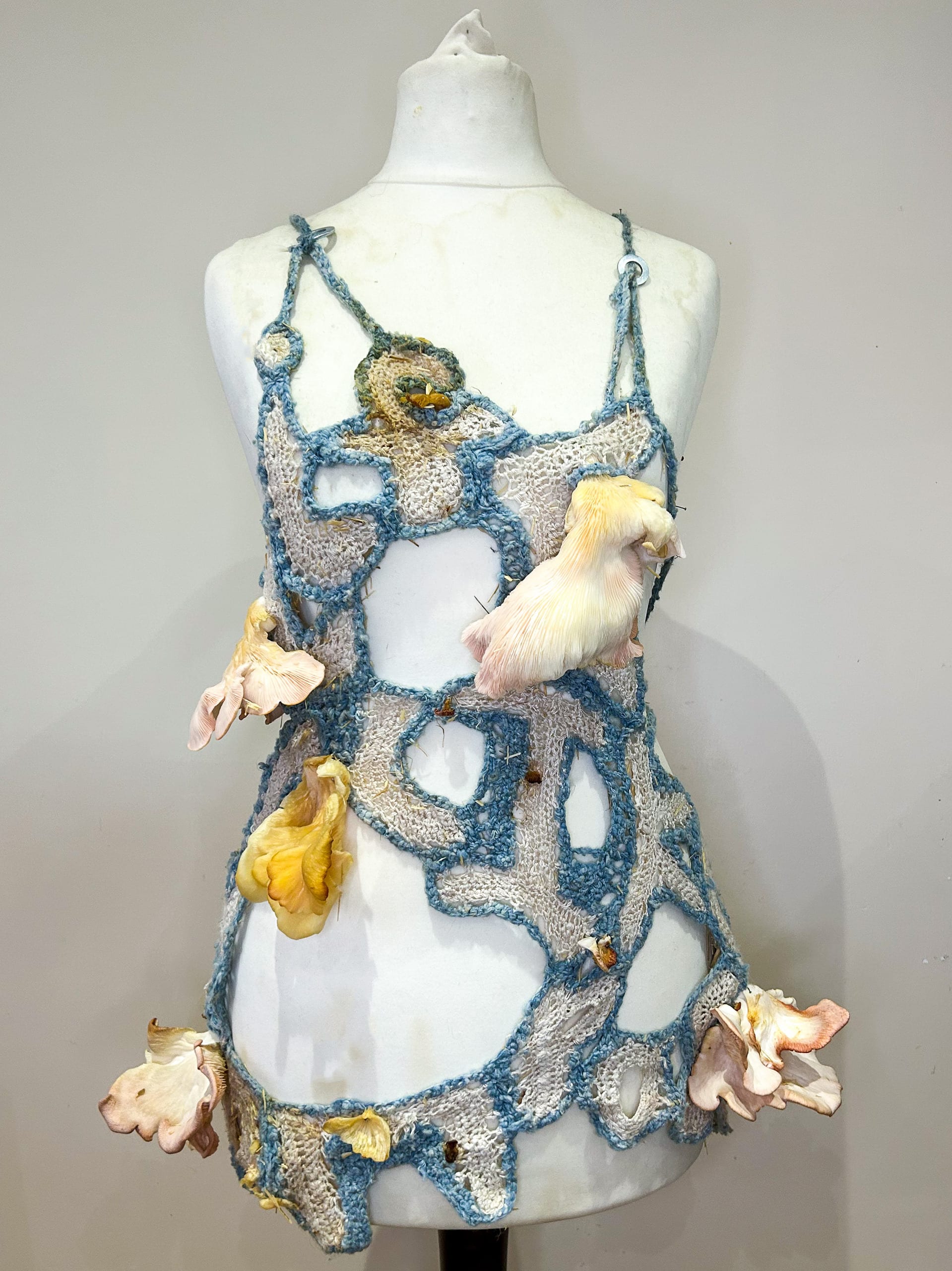 Crocheted letter tie top on dress stand with pink oyster mushrooms growing sporadically out of it.