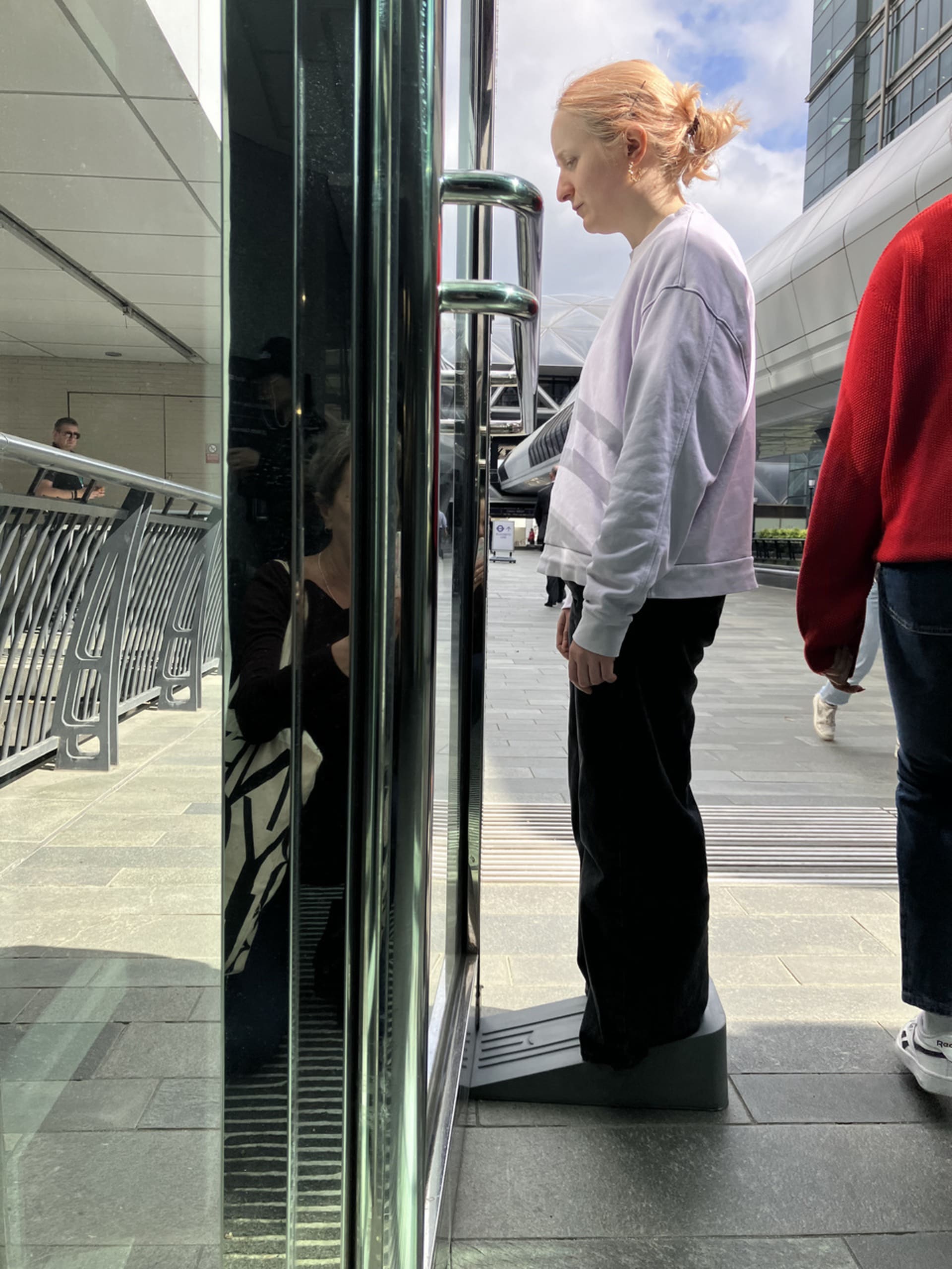 Lili wears a large doorstop shoe to hold open a door for the public in canary warf.