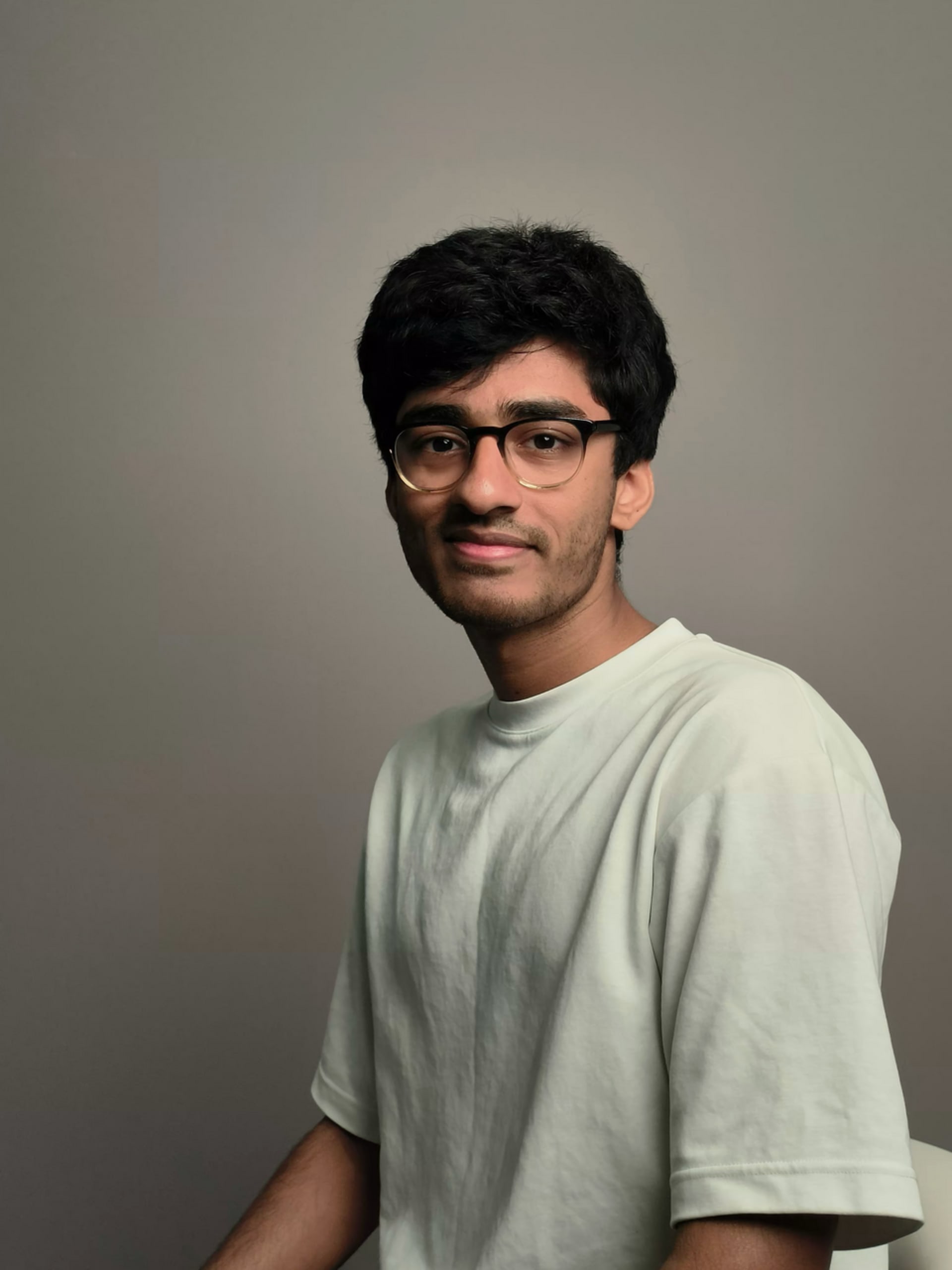 Hunaid sitting on a chair in front of a solid background wearing a white tshirt.