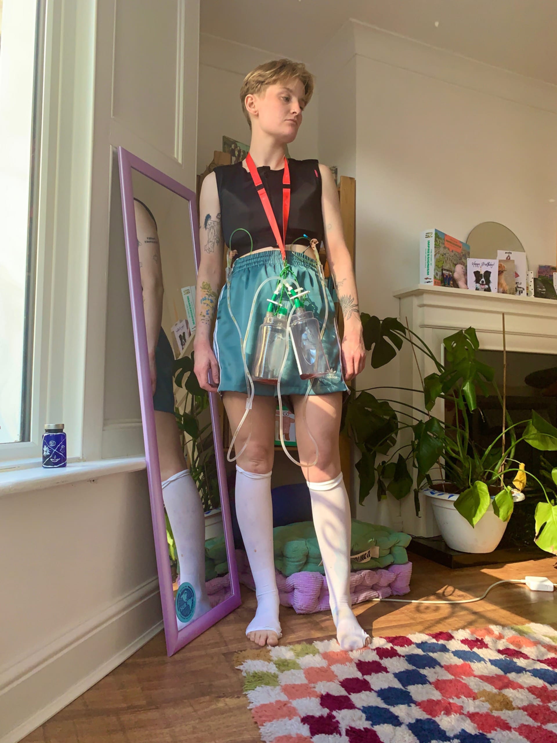 Theo stands in a living room wearing chest drains and compression socks and vest, their head is turned to the side