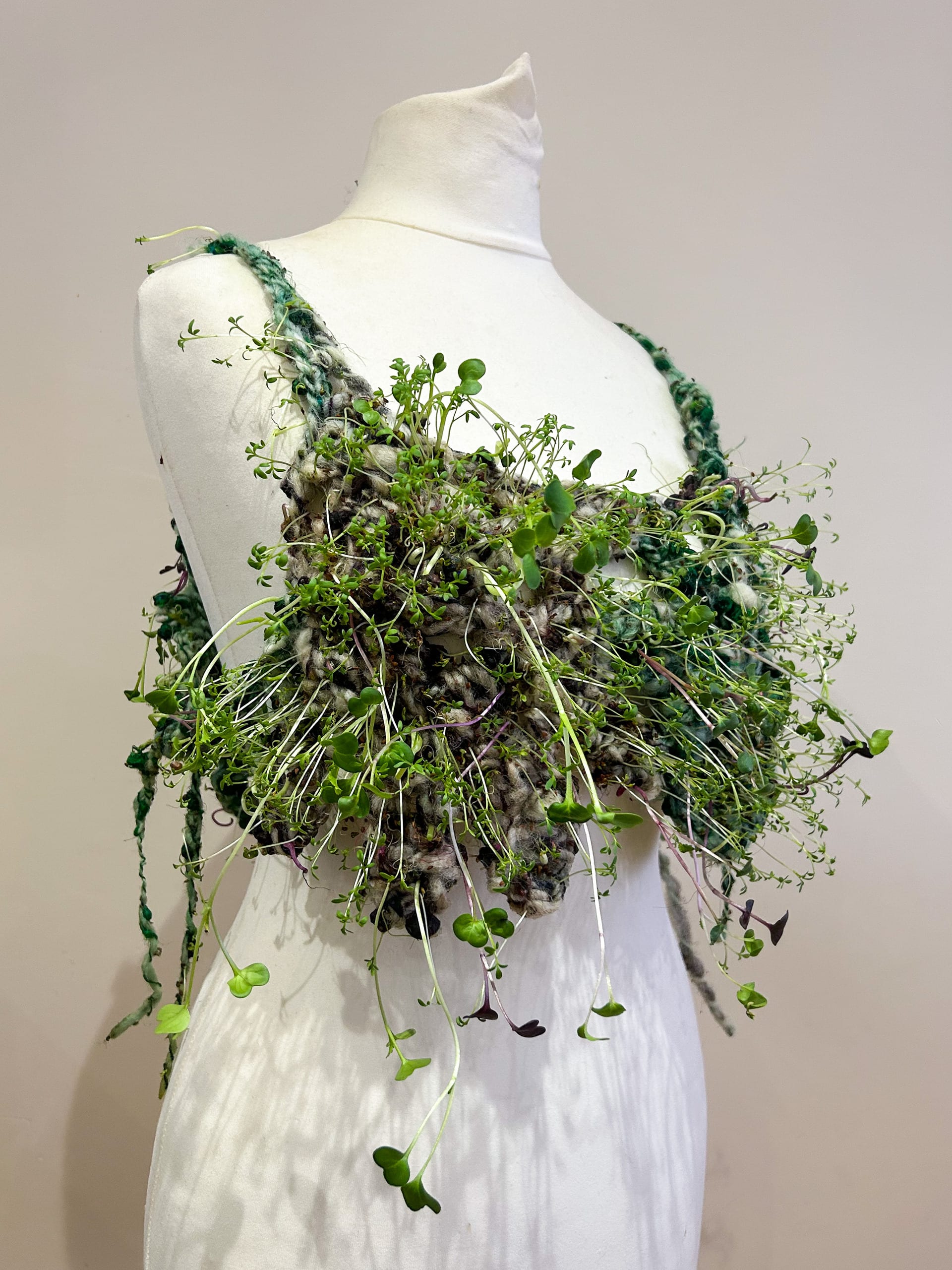 crocheted growing plant top on dress stand, with ties at side against plain wall. stand faced forward but angled. 