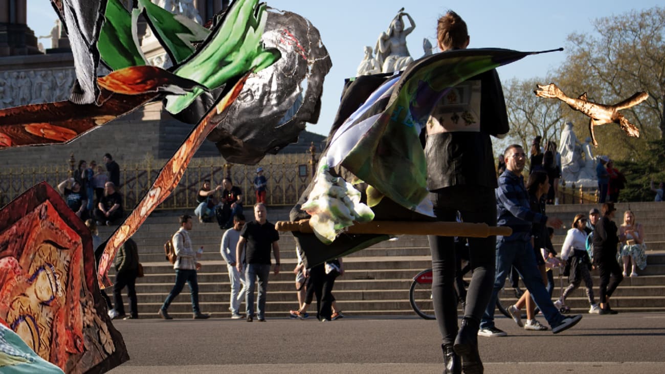 With fabrics draped from the back, a body wearing a patchwork black jacket is walking towards a public sculptures in Hyde Park.