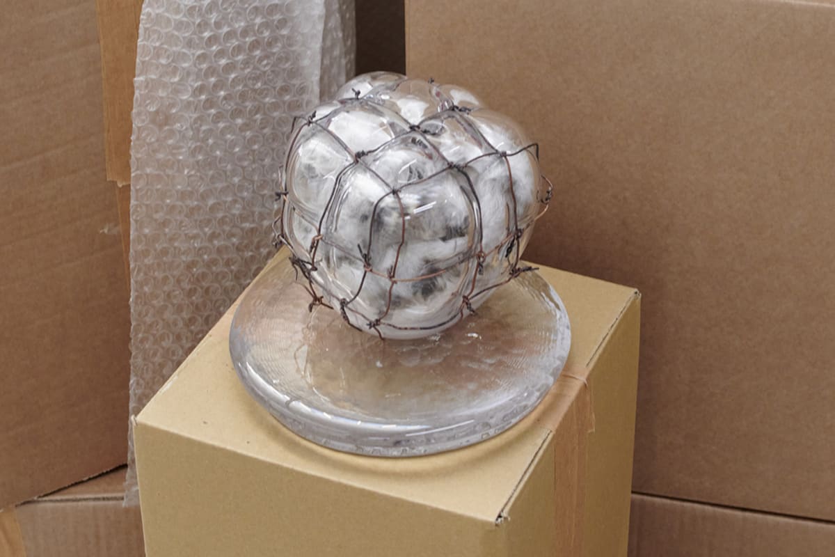 an orb of glass constricted by wire cage is filled with grey feathers and sits on two circles of glass