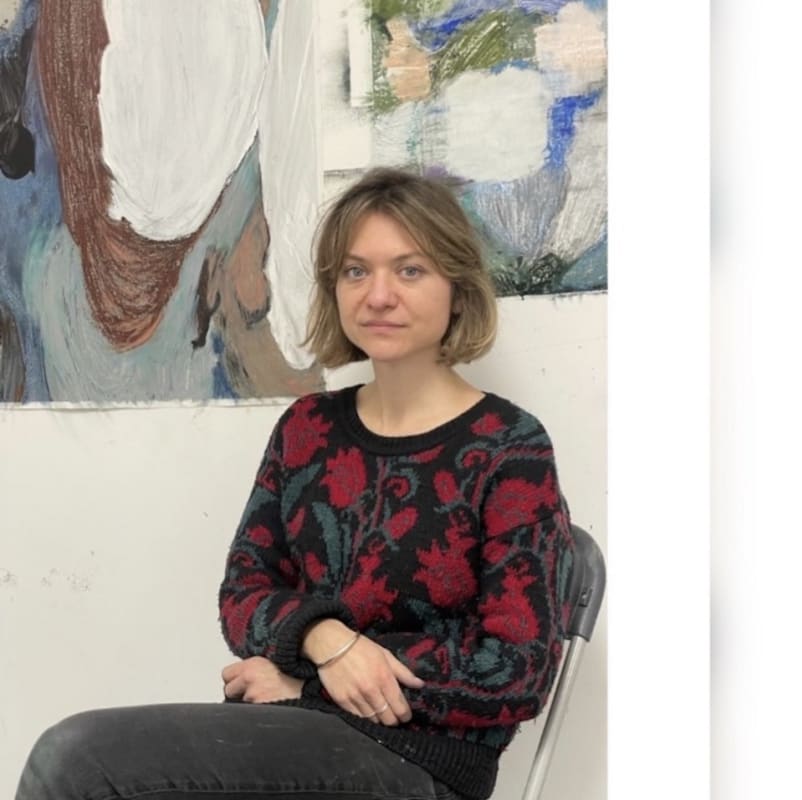 A portrait of the artist seated against the studio wall. She has a mid-brown bob and is wearing black jeans and a patterned top.