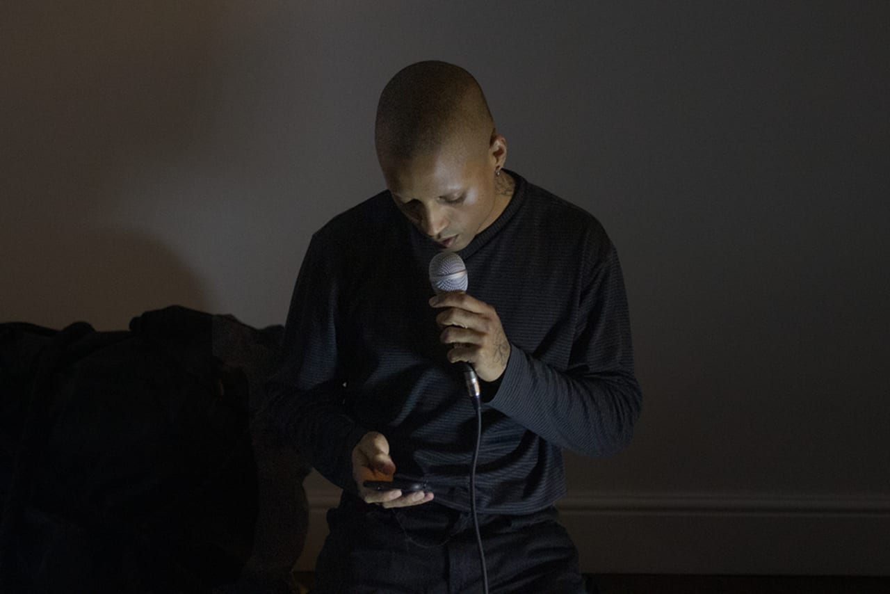 Josh Woolford sat on the floor reading a poem from their iPhone while holding a mic up to their mouth