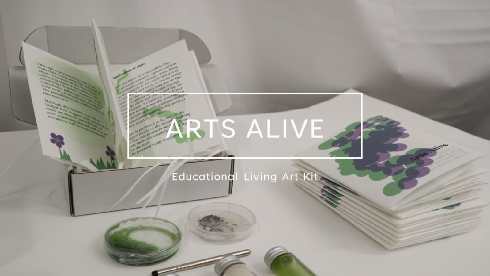 An algae art kit, with two  petri dishes, one green and one dark purple, next to a box with a educational booklet.