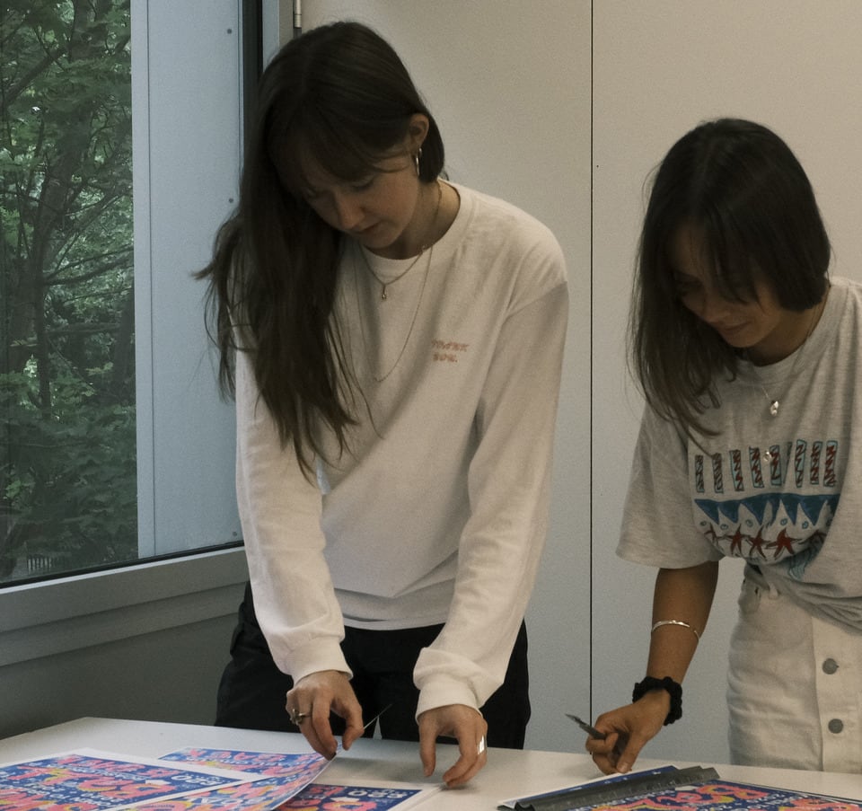 A picture of two women with long hair and fringes sorting through posters. Anya is the woman on the left.