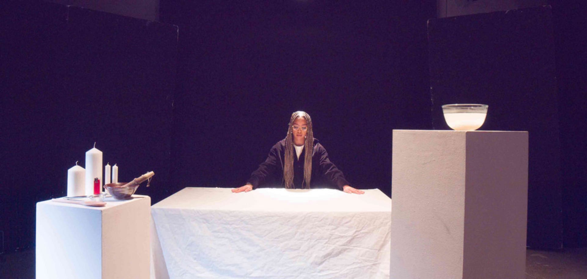 Wide shot of light brown woman with long braids and her hands on a white table; two plinths in front with props on them