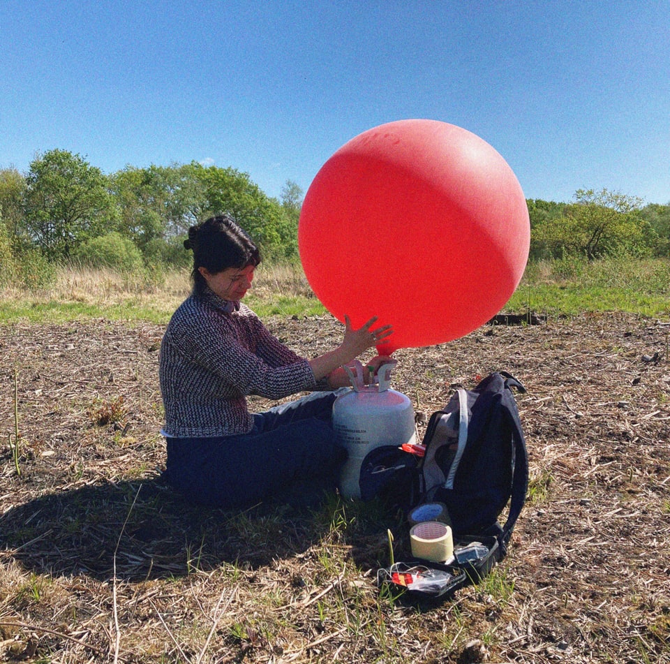 Phoebe sitting on Wimbledon Common inflating a giant red weather balloon.