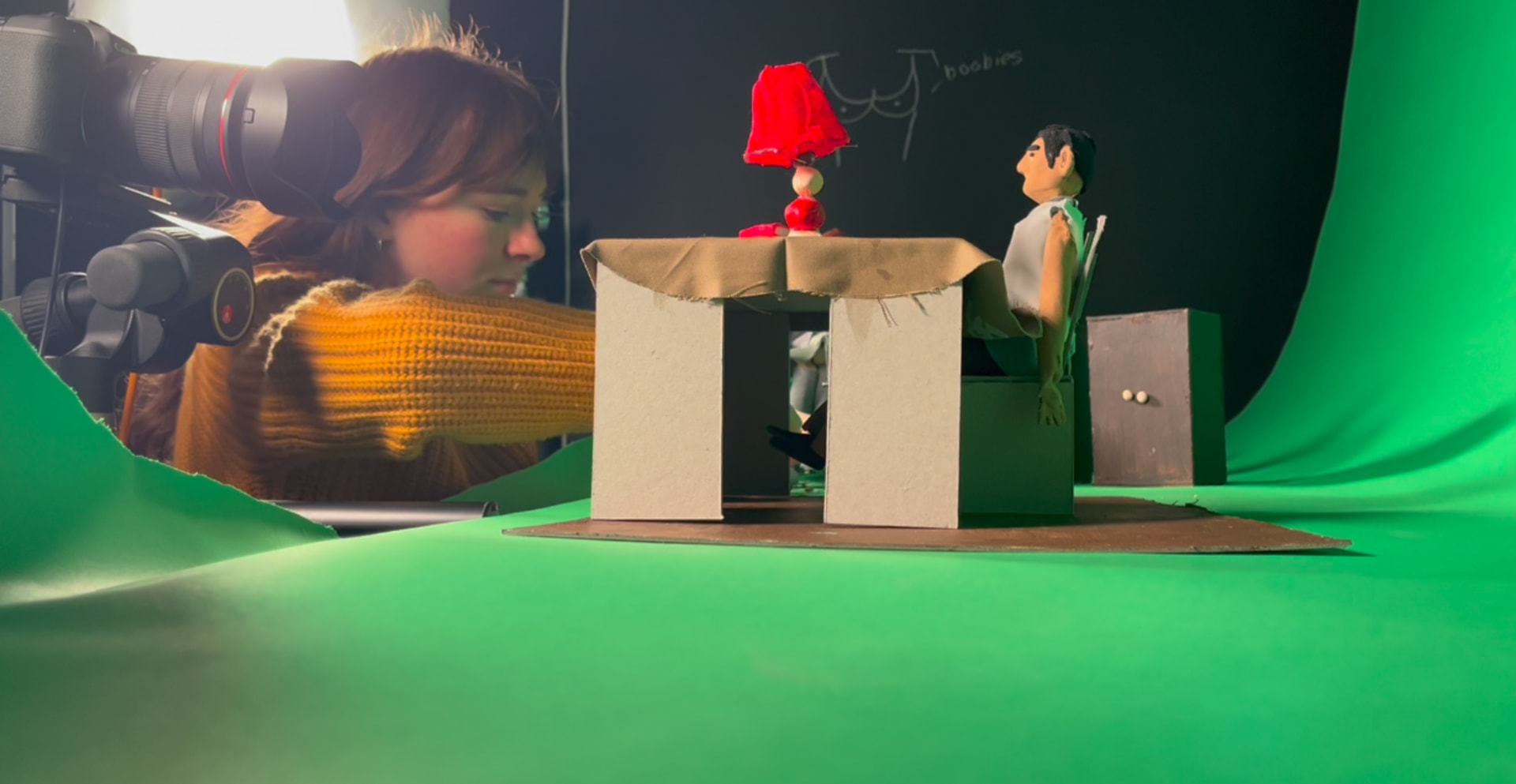 Woman in a capture studio, puppet in the foreground sitting at a table, on a green screen floor and background