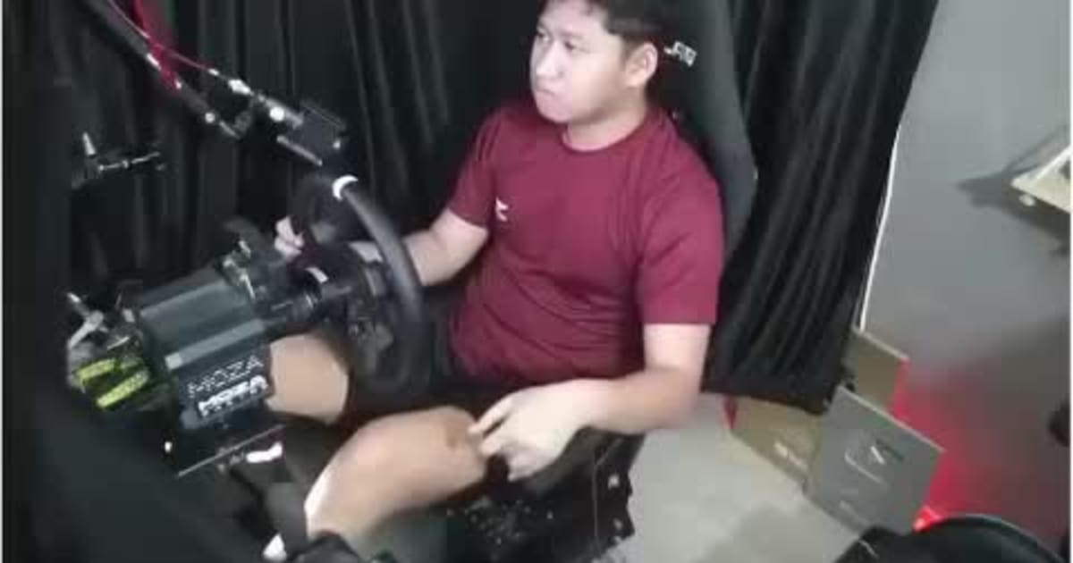 Un joven juega concentrado en un simulador de conducción con volante, mientras en la pantalla se ve un auto esquivando el tráfico en una autopista.