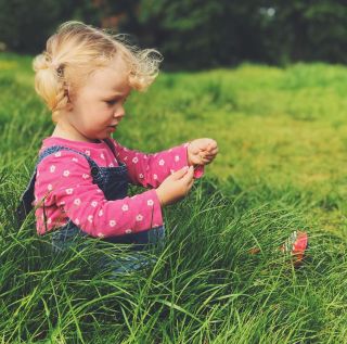 A little girl sat in the gradd wearing a bright pink flowery t-shirt and dungarees