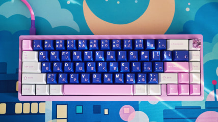 A pink/purple 65% keyboard with blue, pink, and white keycaps against a deskmat inspired by the cityscape background of Sailor Moon.